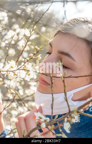 Nahaufnahme Porträt von zartem Mädchen mit geschlossenen Augen unter einem blühenden Aprikosenbaum mit einer Maske aus dem Coronavirus. Stockfoto