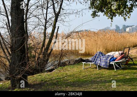 Ein alter Herr, der an einem sonnigen Wintertag auf einem Kinderbett am See in der Nähe eines Schilfs schläft, Gardasee, Bardolino, Verona, Venetien, Italien Stockfoto