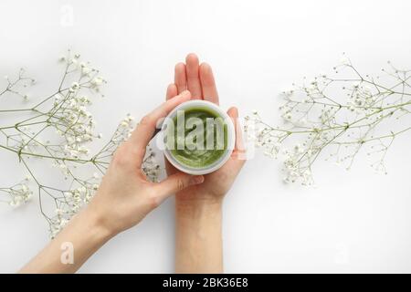 Junge weibliche Hände halten ein Glas grüne natürliche Creme für Gesicht oder Körper. Bio natürliche Hautpflege-Produkte und Blume auf weiß. Verpackung von Lotion oder c Stockfoto