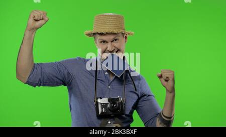 Portrait des Mannes Mode touristischen Fotografen mit Pass in seinem Mund feiert und tanzt. Schöner Mann im Urlaub in blauem Hemd und Hut mit Retro-Kamera. Chroma-Taste. Grüner Bildschirm Stockfoto