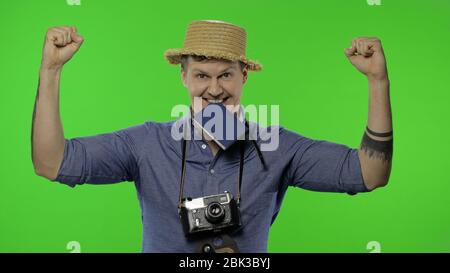 Portrait des Mannes Mode touristischen Fotografen mit Pass in seinem Mund feiert und tanzt. Schöner Mann im Urlaub in blauem Hemd und Hut mit Retro-Kamera. Chroma-Taste. Grüner Bildschirm Stockfoto