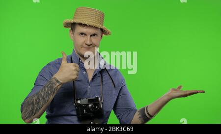 Porträt des Mannes Mode touristischen Fotografen zeigt auf etwas mit seiner Hand. Schöner Mann im Urlaub in blauem Hemd und Hut mit Retro-Kamera. Platz für Ihr Logo. Chroma-Taste. Grüner Bildschirm Stockfoto
