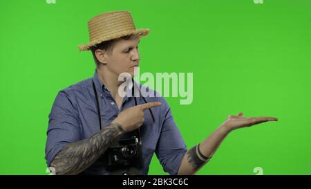 Porträt des Mannes Mode touristischen Fotografen zeigt auf etwas mit seiner Hand. Schöner Mann im Urlaub in blauem Hemd und Hut mit Retro-Kamera. Platz für Ihr Logo. Chroma-Taste. Grüner Bildschirm Stockfoto