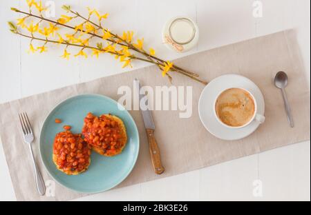 Tisch mit hausgemachten würzigen gebackenen Bohnen mit Speck und Kartoffelbröseln, Tasse Kaffee, Glas Sahne, mit wenigen Zweigen von goldenem Regen verziert. Englisch Stockfoto