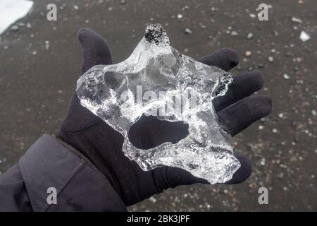 Nahaufnahme eines der vielen Eisstücke, die in einer Hand mit Handschuhen gehalten werden, Jökulsárlón Diamantenstrand, Südisland. Stockfoto
