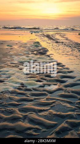 Strand mit Wellen- und Wellenmuster im Sand ist teilweise mit Schaum bedeckt. Die Sonne geht im Westen unter. Stockfoto