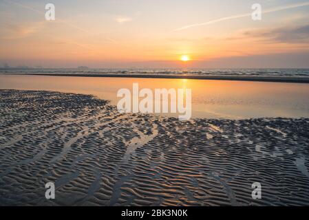 Die Sonne geht hinter einer Wolkenbank über der Nordsee unter. Schöne Farbspiegelung im nassen Sand. Stockfoto