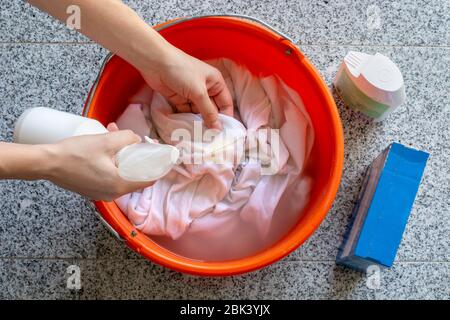 Damenhände waschen schmutzige weiße Hemden mit einem Fleckenentferner aus einer Sprühwaschmittelflasche für gelbe Schweißflecken in einem Eimer Stockfoto
