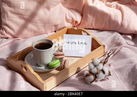 Ein Morgengetränk mit Makronen auf einem Holztablett im Bett. Romantisches Frühstück in einem Bett. Brief Bleib zu Hause. Stockfoto