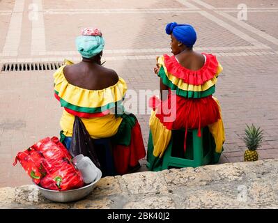 Cartagena, Kolumbien, 1. August 2019: Zwei typische Obsthändler palenqueras mit bunten Kleidern sitzen ruhen von hinten Stockfoto