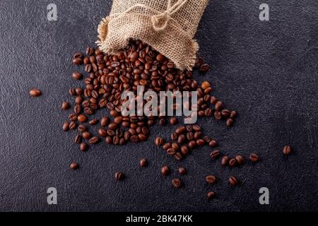 Geröstete, aromatische Kaffeebohnen, die aus einem Leinenbeutel auf einer dunklen Oberfläche bestreut wurden. Das Konzept der Tradition des Morgenkaffees. Draufsicht. Flaches Lay Stockfoto
