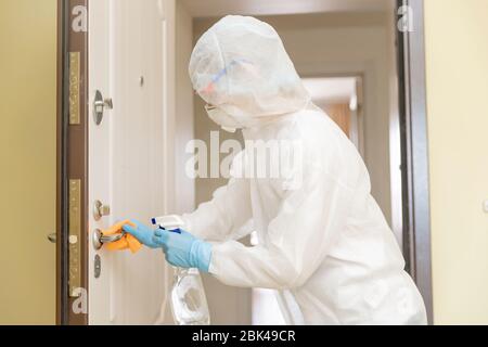 Frau Arbeiter desinfiziert Türgriffe des Hauses von der Infektion mit Viren und Mikroben in biochemischen Anzug. Coronavirus-Schutzkonzept Stockfoto