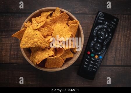 Chips in einer Holzschüssel neben der Fernbedienung des Fernsehers vor einem verbrannten Holzhintergrund. Draufsicht. Nahaufnahme. Getönte. Stockfoto