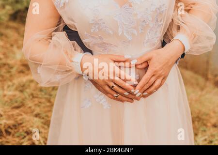 Zwei frisch vermählte Vermählten halten die Hände an einem herzförmigen Bauch. Hochzeitstag. Stockfoto