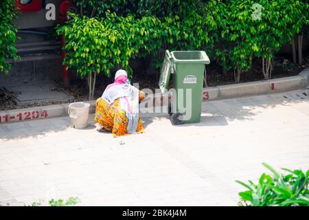 Alte Dame in Saree und Maske Reinigung Gehwege und Straßen mit einem Holzbesen und Müllabfuhr in eine grüne bewegliche Mülleimer Stockfoto