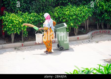 Alte Dame in Saree und Maske Reinigung Gehwege und Straßen mit einem Holzbesen und Müllabfuhr in eine grüne bewegliche Mülleimer Stockfoto