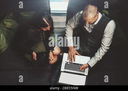 Blick von oben auf zwei Geschäftspartner, die in einem Coworking-Bereich auf gemütlichen Sofas nebeneinander sitzen: Ein Mann Unternehmer mit Laptop und sein f Stockfoto