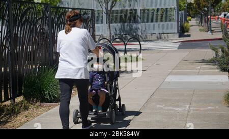Asiatisch-amerikanische Mutter, die während der COVID19-Stilllegung ihr Baby oder Kleinkind im Kinderwagen trägt, beide tragen eine Maske, während sie in Emeryville, CA, USA spazieren. Stockfoto