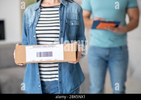 Weibliche Hände halten braunen Karton, männlicher Kurier hinter ihr mit Clip-Ordner Stockfoto