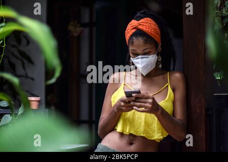 Junge Frau, die während der Sperrung des Coronavirus in Kolumbien eine Gesichtsmaske trägt und ihr Mobiltelefon benutzt Stockfoto