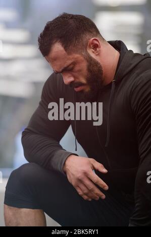 Sport Motivation Konzept der junge bärtige starke Mann Athlet in Sportbekleidung Hoodie lehnt sich an Knie während Pause im Training Stockfoto