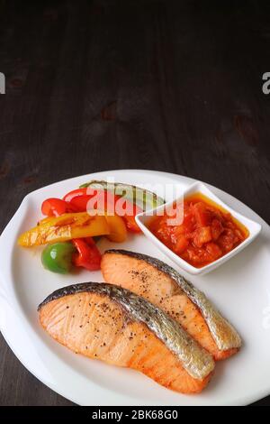 Teller mit in der Pfanne angebratenen Lachssteaks und gegrilltem farbenfrohem Gemüse auf einem dunkelbraunen Holztisch Stockfoto