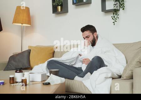 Kranker junger Mann mit Grippe oder Coronavirus sitzt auf der Couch im Wohnzimmer während husten in Decke während Selbstisolation zu Hause Stockfoto