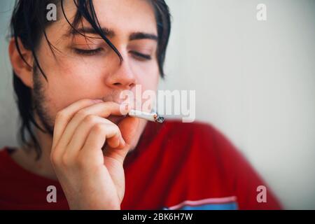 Ein junger, schöner Kerl in einem roten T-Shirt mit nassen Haaren nach einer Dusche genießt das Rauchen einer Zigarette am frühen Morgen. Stockfoto
