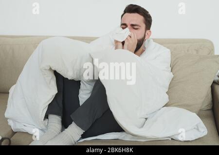 Junger Mann mit Grippe oder Kovid19 hustet in Taschentuch oder Papiertuch, während er während der Selbstisolation auf der Couch im Wohnzimmer sitzt Stockfoto