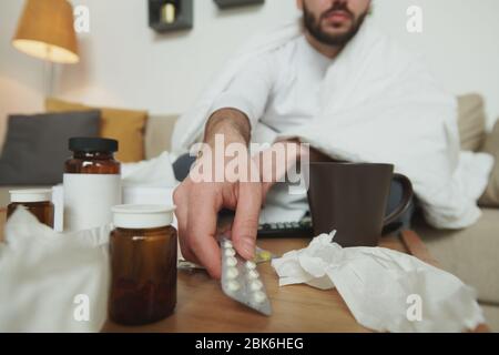 Hand des kranken jungen Mannes, der Blister mit Schmerzmitteln oder schleimlösenden Medikamente vom Tisch nimmt, während er während der Selbstisolierung zu Hause auf dem Bett oder der Couch sitzt Stockfoto