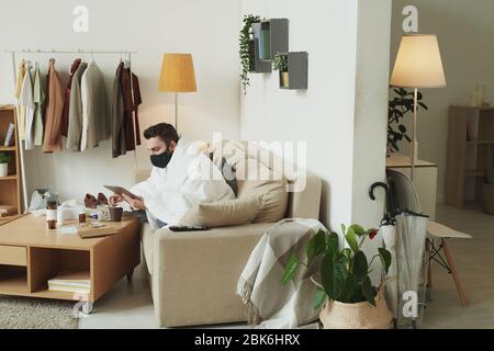 Kranker junger Mann in schwarzer Maske sitzt auf der Couch und mit Touchpad während der Arbeit entfernt in der häuslichen Umgebung während der Selbstisolierung Stockfoto