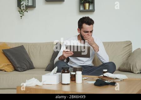 Sehr kranker junger Mann mit digitalem Tablet, der auf der Couch sitzt und seinen Mund bedeckt, während er bei der Arbeit im Haus hustet Stockfoto