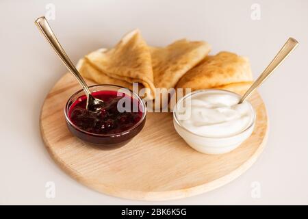 Zwei kleine Glasschüsseln mit hausgemachter Kirschmarmelade und Sauerrahm und drei heißen, appetitlich gefalteten Pfannkuchen auf Holzbrett Stockfoto