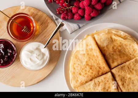 Flacher Grundriss mit vier gefalteten Pfannkuchen auf Teller, frischen Himbeeren und roten Johannisbeeren, kleinen Glasschüsseln mit Honig, Kirschmarmelade und Sauerrahm an Bord Stockfoto