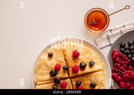 Flatlay von Küchentisch mit appetitlichen hausgemachten Pfannkuchen mit frischen Beeren auf dem Teller, kleine Glasschüssel mit Honig und gefaltete Serviette Stockfoto