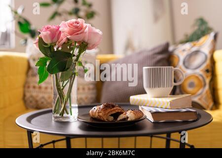 Ein Bündel frischer rosa Rosen in einem Glas Wasser, hausgemachtes Croissant, eine Tasse Tee oder Kaffee und zwei Bücher auf einem Tablett auf dem Hintergrund der Couch Stockfoto