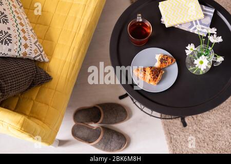 Flache Anordnung von Pantoffeln auf dem Boden zwischen gelben Couch mit Kissen und kleinem Tisch mit Büchern, Glas Tee, Croissants und Blumen in Krug Stockfoto