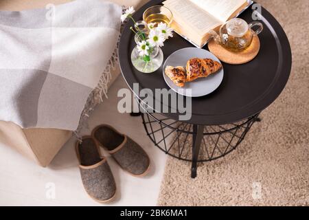 Kleiner Tisch mit offenem Buch, Blumenstrauß, knusprigen Croissants und grünem Tee, der am Sessel mit Plaid und Pantoffeln auf dem Boden steht Stockfoto