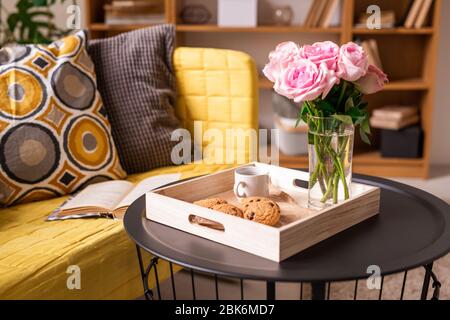 Inneneinrichtung mit Kissen und offenem Buch auf Couch und kleinem Tisch mit Plätzchen, Tasse Kaffee und Bündel rosa Rosen in Holzkiste Stockfoto