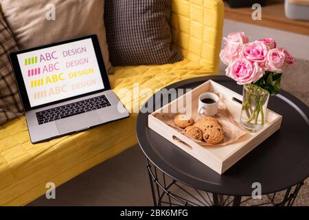 Laptop und Kissen auf der gelben Couch von einem kleinen Tisch mit Holzkiste mit Bündel rosa Rosen, Getränk und leckere Kekse Stockfoto