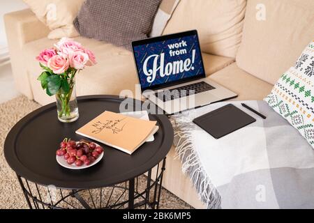 Kleiner runder Tisch mit Rosenstrauß, frischen roten Trauben auf Teller und Skizzenbuch, das von der Couch mit Laptop und Pad des Designers steht Stockfoto