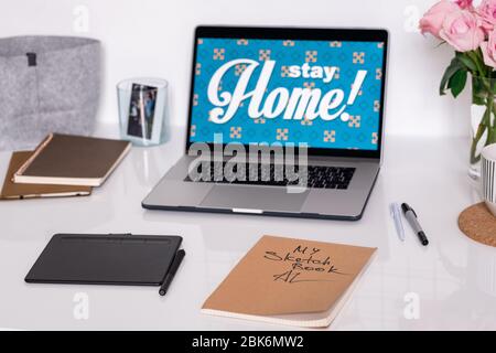 Laptop mit Stay Home Ansage auf dem Display, Pad mit Stylus, Skizzenbuch, Stifte, Notizbücher und Rosen auf dem Arbeitsplatz des Designers Stockfoto