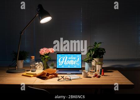 Arbeitsplatz durch Fenster in einem dunklen Raum mit Laptop, Papiersack mit Croissant, Blumen, Getränken, Kosmetikartikeln, Büchern auf dem Tisch und Lampe oben Stockfoto