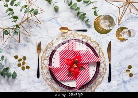 Weihnachtstisch mit roter Serviette, Weihnachtsstern, goldenen Utensilien und Eukalyptusblättern auf Marmorgrund. Flach auf Tisch mit goldenem Besteck gelegt, Stockfoto