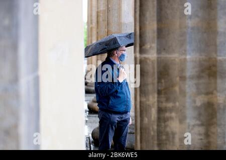 Berlin, Deutschland. Mai 2020. Ein Mann mit Mund-Nase-Schutz steht mit einem Regenschirm zwischen den Säulen des Brandenburger Tors. Quelle: Christoph Soeder/dpa/Alamy Live News Stockfoto