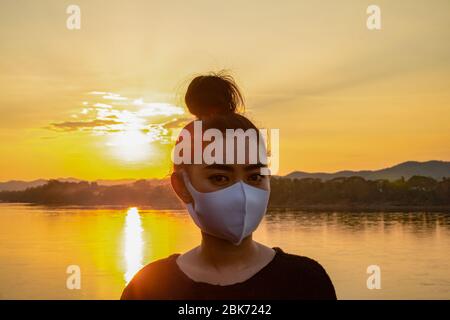 Nahaufnahme junge Asienfrau, die eine Fack Maske aufsetzt, um vor luftgetragenen Atemwegserkrankungen zu schützen, wie die Grippe-covid-19 PM2.5 Staub und Smog bei Sonnenuntergang backg Stockfoto