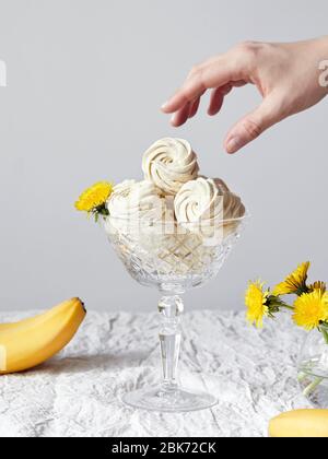 Gelb süß hausgemachte Zephyr oder Marshmallow aus Banane in Glasvase auf weißem Hintergrund. Frau will eine nehmen. In der Nähe von frischen Bananen und Löwenzahn fl Stockfoto