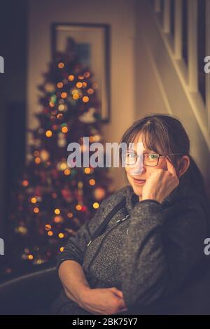 Nahaufnahme einer isolierten Dame, die im Stuhl sitzt und sich vor einem festlich geschmückten Weihnachtsbaum in Großbritannien nach dem Kochen und Essen des Weihnachtsessens entspannt. Stockfoto