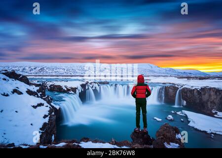 Godafoss Wasserfall bei Sonnenuntergang im Winter, Island. Kerl in roter Jacke sieht Godafoss Wasserfall. Stockfoto