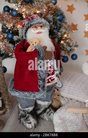 Weihnachtsmann Puppe, große Weihnachtsstandfigur hält niedlichen Teddybär, vor einem beleuchteten Weihnachtsbaum positioniert, festliche Dekoration für Feiertage Stockfoto